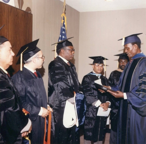 University officer presenting a bound honour to a candidate in doctoral regalia before assembled faculty