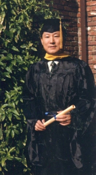 Graduate in cap and gown holding a diploma scroll beside a garden wall