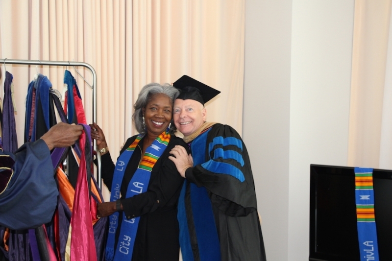 Two members of the university community smiling beside a rack of academic hoods