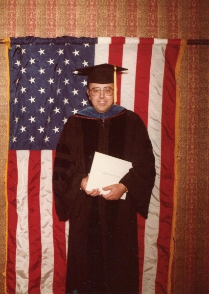 Man in academic regalia holding a diploma before an American flag