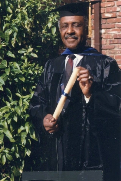Graduate in black regalia with blue hood holding a diploma scroll outdoors