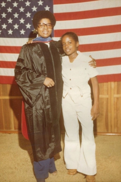 Graduate in academic regalia standing with a young family member before an American flag