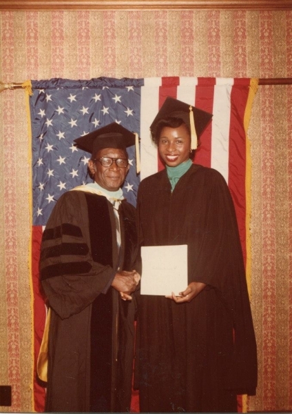 Faculty member in doctoral regalia shaking hands with a graduate holding a diploma before an American flag