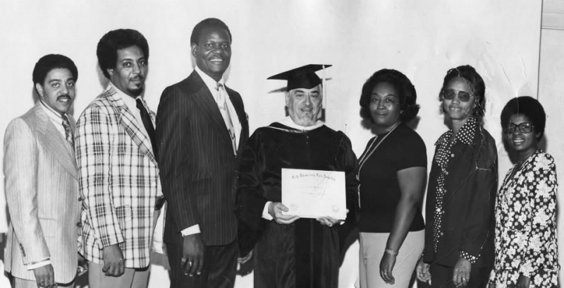 Honouree in cap and gown holding a certificate surrounded by family and guests