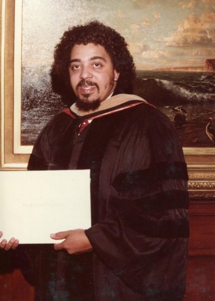 Graduate in doctoral regalia holding a diploma in front of an oil painting