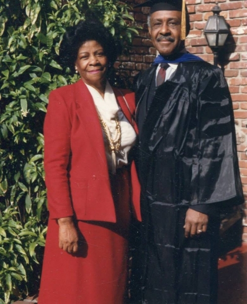 Graduate in cap and gown standing with a family member outside a brick building
