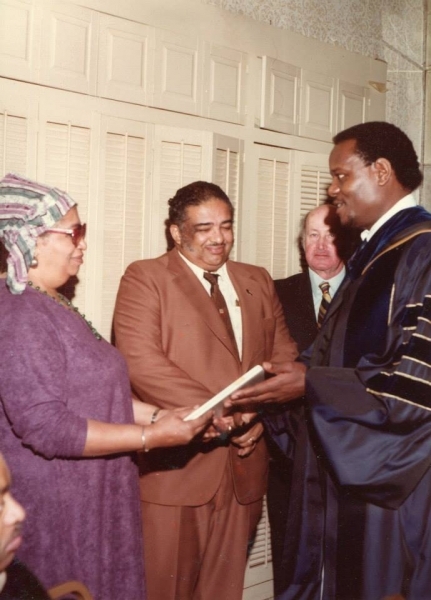 University officer in doctoral regalia handing a diploma to an honouree and his wife