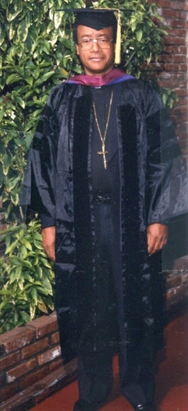Portrait of a doctoral graduate in full regalia beside a brick wall
