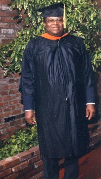 Graduate in cap and gown standing outdoors beside a brick wall