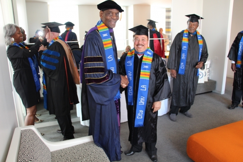 Two university officers shaking hands in regalia as faculty gather nearby
