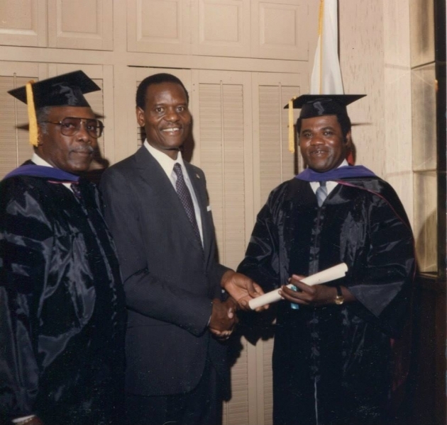 University officer congratulating a graduate holding a diploma scroll beside a faculty member