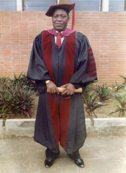 Graduate in crimson and black doctoral regalia holding a diploma outdoors