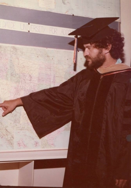 Graduate in regalia pointing to a world map on a classroom wall