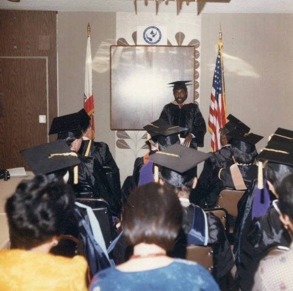 Officer in regalia addressing seated graduates beneath the university seal and flags