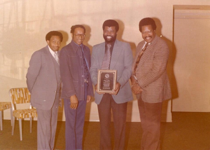 Four men standing together as an honouree holds an engraved award plaque